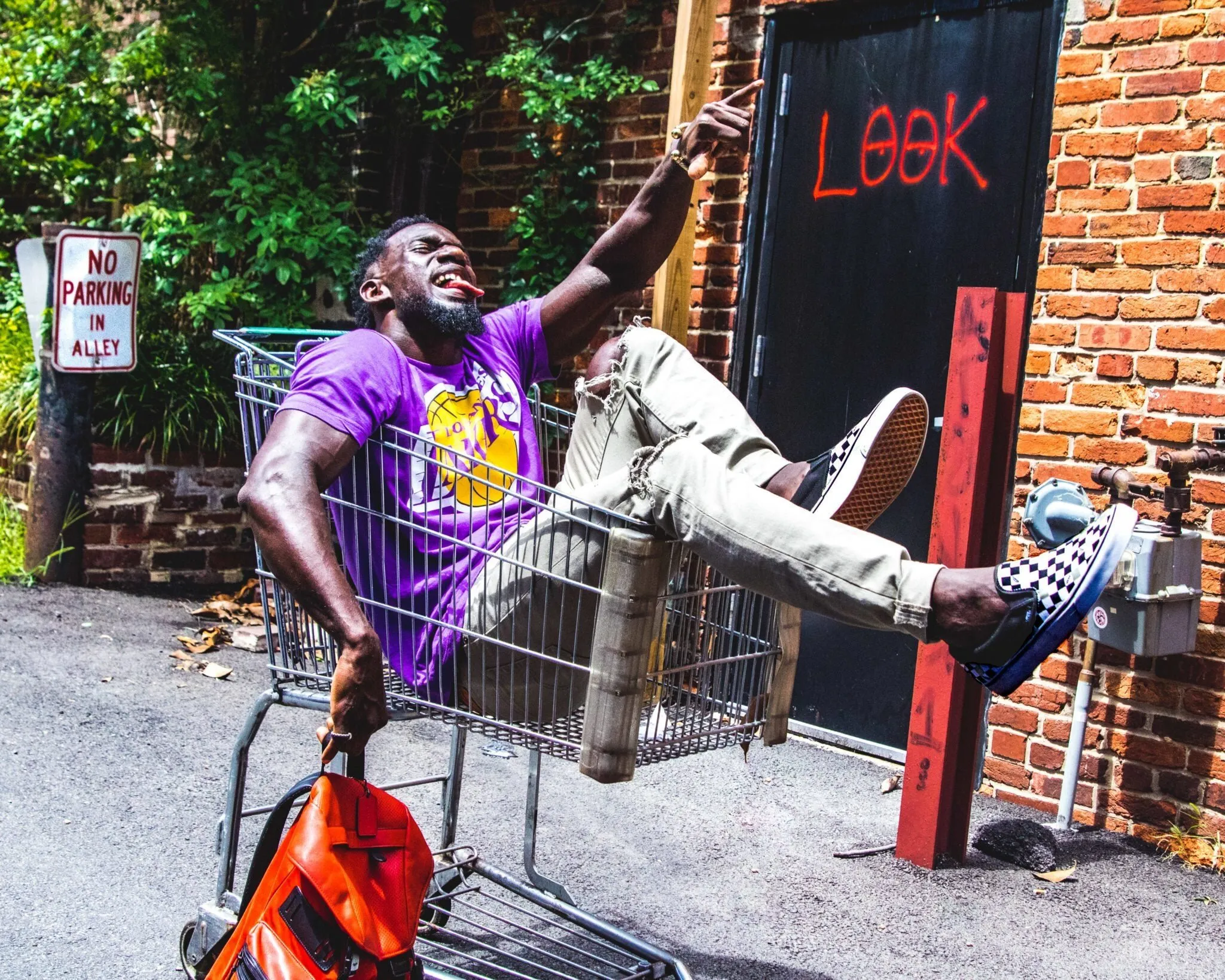 A young man riding a shopping cart down an alley, arms out, past bright street art